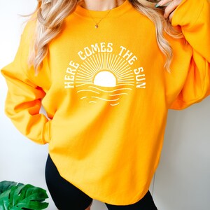Puede incluir: Una sudadera amarilla con un gr&aacute;fico blanco de un sol saliendo sobre el oc&eacute;ano y el texto "Here Comes The Sun".