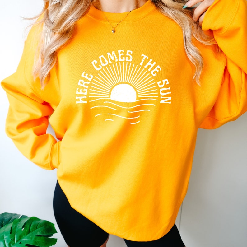 Puede incluir: Una sudadera amarilla con un gr&aacute;fico blanco de un sol saliendo sobre el oc&eacute;ano y el texto "Here Comes The Sun".