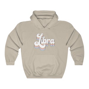 K&ouml;nnte beinhalten: Ein hellbeiger Hoodie mit einem wei&szlig;en Retro-&bdquo;Libra&ldquo;-Motiv in Regenbogenfarben.