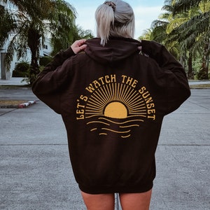 Puede incluir: Una sudadera con capucha negra con un gráfico amarillo en la espalda que dice "Let's watch the sunset" con un sol y olas estilizados.