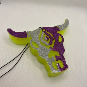 May include: A decorative air freshener shaped like a bull skull. The skull is lime green with silver and purple glitter accents. The horns and details are silver glitter, and the design is purple glitter. Black string is attached for hanging.
