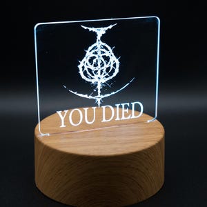 May include: A white LED light with a wooden base. The light features a graphic of a symbol and the text "YOU DIED".