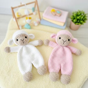 以下が含まれることがあります： 手作りの編みぐるみの子羊人形が2つ。1つは白、もう1つはピンクです。どちらもベージュの顔、耳、手、足があります。黄色い毛布の上に横たわっています。背景には、木製のベビージム、パステルカラーの毛布の山、小さな鉢植えの植物が含まれています。