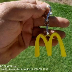 Op de afbeelding: Een gele plastic sleutelhanger in de vorm van het gouden McDonald's-booglogo. De sleutelhanger is bevestigd aan een zilveren metalen ring en een zilveren metalen ketting.