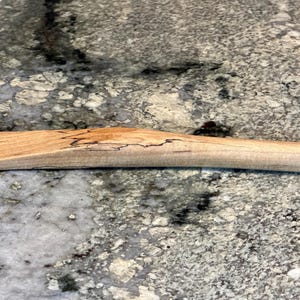 May include: A hand-carved wooden spreader with a flat, wide blade and a rounded handle. The wood features a light tan color with natural grain patterns and dark, irregular spalted markings running along the center of the handle.