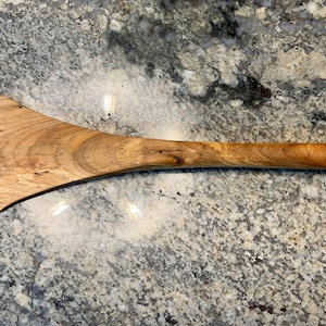 May include: A handcrafted wooden spatula featuring a wide, flat head that tapers into a rounded, ergonomic handle. The utensil displays a natural light brown wood grain with visible knots and organic patterns throughout its smooth, polished surface.
