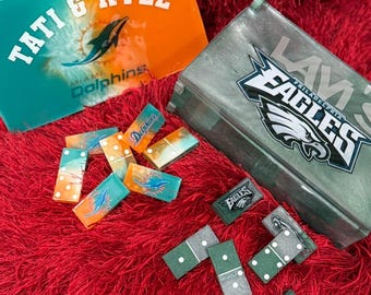 May include: Two custom domino sets with matching boxes. One set features the Miami Dolphins logo and the names "Tati & Kyle" on an orange and teal box. The other set has the Philadelphia Eagles logo on a gray box.