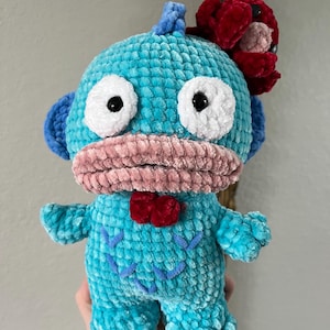May include: A handmade turquoise plush toy with large white eyes and a pink mouth. It has a red bow tie and a red flower on its head. The toy is crocheted with a soft, textured yarn.