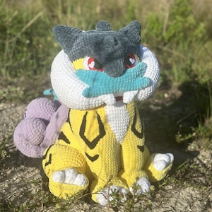 Pode incluir: Um peluche de croché feito à mão com corpo amarelo e riscas pretas, cabeça cinzento-escura e colarinho branco. A criatura tem olhos vermelhos, focinho azul-petróleo e uma cauda roxa bulbosa. Está sentada com patas brancas e costuras.