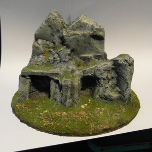 May include: A miniature, hand-painted model of a stone structure, possibly a ruin or a fantasy landscape element. The model features a grey, textured stone appearance with patches of green moss and a grassy base. The structure has archways and a layered design.