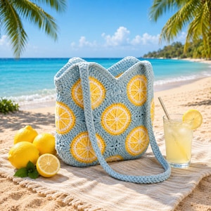 May include: A light blue crochet tote bag featuring yellow and white lemon slice patterns. The bag has a matching blue strap and is placed on a beach towel alongside lemons and a glass of lemonade. The background shows a beach and ocean.
