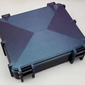 May include: A square, dark blue case with a geometric design on the lid. The case has a textured surface and black hinges and clasps. The case is designed to protect its contents.