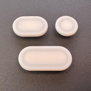 May include: Three off-white, oval and circular shaped objects. The objects have a slightly raised edge and a smooth, matte finish. The shapes are arranged on a dark gray surface.