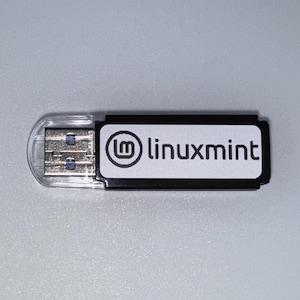 May include: A USB flash drive with a clear plastic cover and a black and white label. The label features the Linux Mint logo and the text "linuxmint" in a sans-serif font. The drive is designed for data storage and transfer.