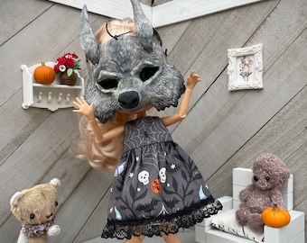 May include: A doll with blonde hair wearing a grey wolf mask and a dark grey sleeveless dress featuring a pattern of white skulls, orange pumpkins, black bats, and spiderwebs, finished with a black lace hem and yellow ankle boots.