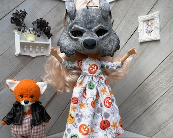 Puede incluir: Una muñeca con una máscara de lobo gris y un vestido blanco con estampado de calabazas. Un zorro de peluche con una chaqueta de cuero y pantalones a cuadros está cerca. Pequeñas calabazas y botas amarillas completan el tema de Halloween.