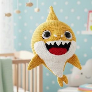 Puede incluir: Un juguete de tiburón tejido a mano en color amarillo con detalles blancos, una boca roja y ojos grandes. El tiburón está sonriendo. El juguete está diseñado para niños y se encuentra en un cuarto de niños con una cuna y un móvil.