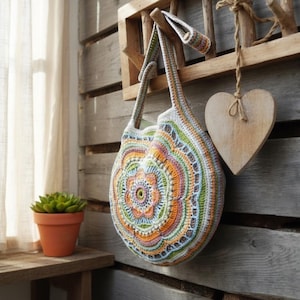 May include: A vibrant, handmade crochet bag featuring a circular floral pattern in orange, green, blue, and white. The bag is suspended from a wooden rack, alongside a wooden heart decoration. A small potted plant is on a wooden shelf.