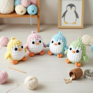 May include: Four colorful crochet penguin toys in yellow, pink, blue, and green. Each penguin has a white belly, black eyes, and orange feet and beak. The penguins are handmade and sit on a white surface.