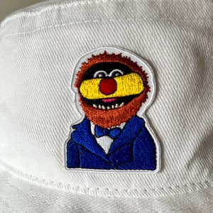 Op de afbeelding: Een witte bucket hat met een geborduurde patch van een personage met een bruine baard, een rode neus, een geel masker en een blauw colbert met een bijpassende blauwe vlinderdas. Het borduurwerk is gedetailleerd met zichtbare stiksels en een witte rand.