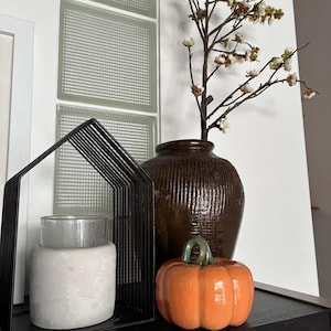 May include: A black metal house-shaped frame containing a white cylindrical candle holder, a dark brown ribbed ceramic vase with white flowering branches, and a small orange ceramic pumpkin with a green stem arranged together.