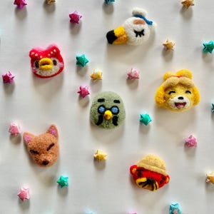 Op de afbeelding: Een verzameling kleurrijke vilten karakterkoppen, waaronder een eend, een vos en een vogel, omringd door stervormige decoraties. De karakters zijn in verschillende kleuren, zoals rood, geel en groen, en zijn gerangschikt op een witte achtergrond.