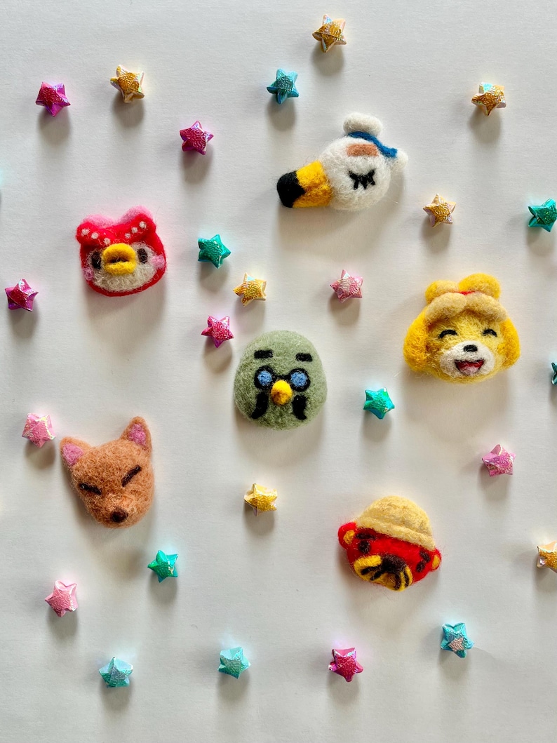 Op de afbeelding: Een verzameling kleurrijke vilten karakterkoppen, waaronder een eend, een vos en een vogel, omringd door stervormige decoraties. De karakters zijn in verschillende kleuren, zoals rood, geel en groen, en zijn gerangschikt op een witte achtergrond.