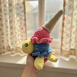 Könnte beinhalten: Handgefertigte Häkelschildkröte mit rosa Eiscreme und einem beigefarbenen Kegel auf dem Rücken. Die Schildkröte ist gelb mit blauen und gelben Akzenten. Das Spielzeug befindet sich auf einer Fensterbank mit Blumenvorhängen im Hintergrund.