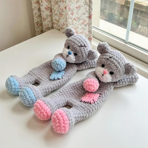 May include: Two handmade gray crochet bear-shaped pillows with blue and pink accents. Each bear has a white muzzle, black stitched eyes, and a small blue or pink pom-pom. The pillows are lying on a white surface.