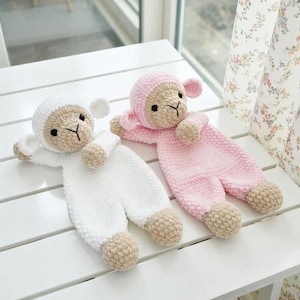 以下が含まれることがあります： 手作りのかぎ針編みの羊のセキュリティブランケット2枚。1つは白、もう1つはピンクです。どちらもベージュの顔、耳、手、足があります。子羊は黒い目とベージュの鼻をしています。ブランケットは白い表面に置かれています。