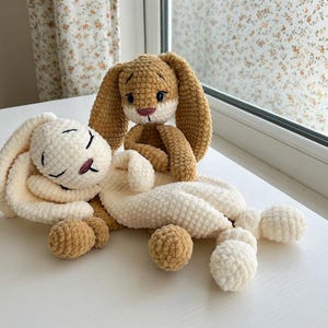 May include: Two handmade crochet bunny rabbits in cream and tan. One bunny is lying down with closed eyes, and the other is sitting up. The bunnies have long ears and are made of soft, textured yarn.