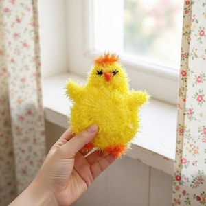 May include: A fluffy yellow chick-shaped item with orange feet and a crest. The chick has black eyelashes and a red beak. The item is held in a person's hand near a window with floral curtains. The item is likely a decorative item or a toy.