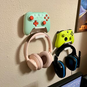 Może przedstawiać: Wiszący uchwyt na słuchawki i kontrolery do gier z dwoma parami słuchawek i dwoma kontrolerami. Jeden kontroler jest zielony, a drugi jasnoniebieski z czerwonymi przyciskami.