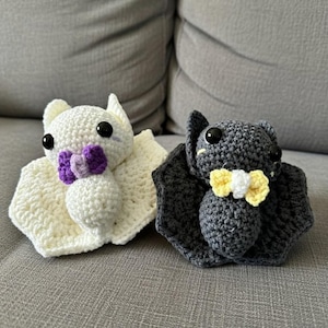 May include: Two crocheted bat plushies. One is white with a purple bow tie, the other is grey with a yellow and white bow tie. Both have large black eyes and are sitting on a grey surface.
