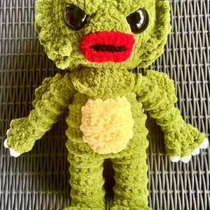 May include: A handmade crochet plush creature featuring a textured olive green body, a pale yellow belly patch, and bright red lips. The figure has large black glossy eyes, a wide head with ruffled edges, and white claws on its hands and feet.