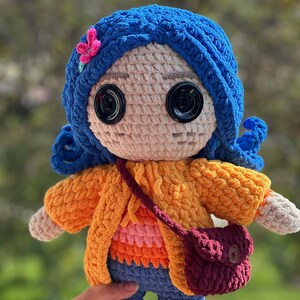 May include: A crocheted doll with blue hair, a pink flower, and a brown purse. The doll is wearing a yellow sweater, an orange and pink shirt, blue pants, and yellow shoes.