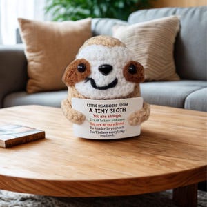 May include: A crocheted sloth figurine holding a white sign with the text "LITTLE REMINDERS FROM A TINY SLOTH". The sloth is beige, brown, and white, with a smiling face. The sign has inspirational text.