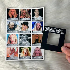 May include: A white card with a grid of 12 photos of Britney Spears making different facial expressions. Each photo has a word below it describing the emotion. The card is next to a black square with a cutout in the middle and the words "CURRENT MOOD..." printed on it.