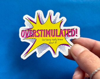 May include: A sticker with the text "OVERSTIMULATED!" in pink, inside a yellow burst shape with purple outlines. The sticker also includes the text "(but being really brave about it)" in yellow. The background is blue.