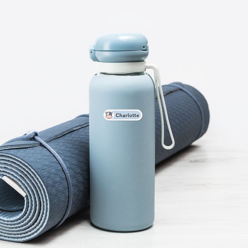 May include: A light blue water bottle with a white strap and a light blue lid. A white label with the name "Charlotte" and a cow graphic is on the bottle. A rolled-up blue yoga mat is in the background.