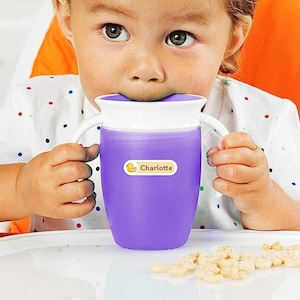 May include: A purple sippy cup with white handles and a white rim. A yellow label with a duck graphic and the name "Charlotte" is affixed to the cup. The cup is being held by a child.