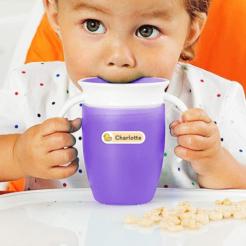 May include: A purple sippy cup with white handles and a white rim. A yellow label with a duck graphic and the name "Charlotte" is affixed to the cup. The cup is being held by a child.