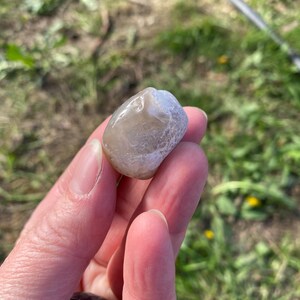 May include: A smooth, rounded, light gray stone with a slightly translucent appearance. The stone has a polished surface and a natural, organic shape. The background is blurred, showing green grass and a hint of a black hose.