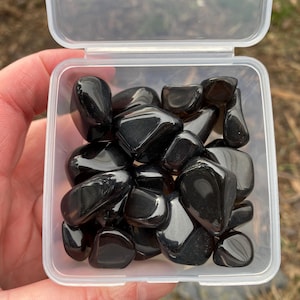 May include: A clear plastic container filled with polished, black obsidian stones. The stones are smooth and reflect light, creating a glossy appearance. The container is held against a blurred outdoor background.