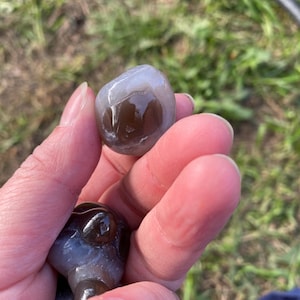 May include: A hand holding two polished stones. The top stone is a rounded shape with a translucent gray and brown pattern. The bottom stone is a dark brown color with a smooth, glossy texture. The background is blurred.