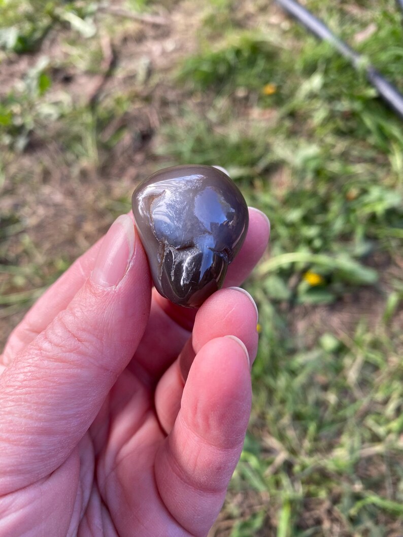 May include: A smooth, polished, gray stone with a glossy surface. The stone has a rounded shape and appears to be held in a person's hand. The background is blurred, showing green grass and a hint of a black object.