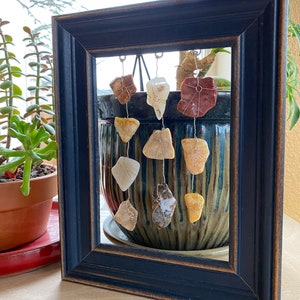 May include: A black picture frame with a glass front displays a collection of colorful, natural stones hanging from thin wires. The stones are various shapes and sizes, and they are arranged in a cascading pattern. The frame is sitting on a wooden surface.