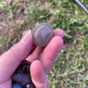 May include: A smooth, gray stone held in a hand, with a glossy surface. The stone has a rounded shape and appears to be polished. Other dark stones are also visible in the hand. The background is blurred, showing green grass.
