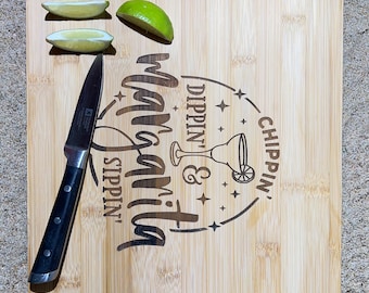 May include: A bamboo chopping board with a knife and lime wedges. The board has an engraved design that says "Margarita Sippin' Dippin' & Chippin'" with a margarita glass and stars.