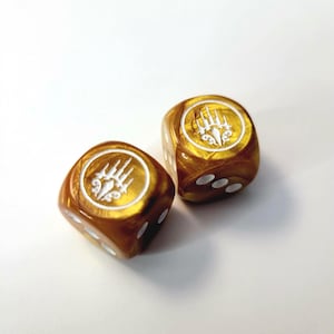 May include: Two gold-colored dice with white pips and a white emblem on each face. The dice have a glossy finish and are set against a white background. The emblem appears to be a stylized design.
